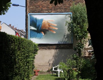 billboard inside a garden. The billboard image is of an arm with a blue jumper and a hand pulling at it. The background is a brick wall and a hedge. The garden is green, with a white deck chair, potted plants and trees