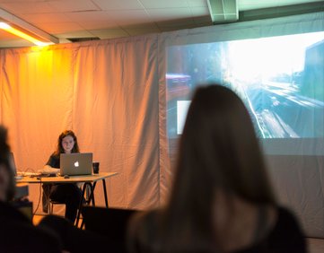 Woman at a table, a screen behind her showing a film, and an audience member looking towards the screen
