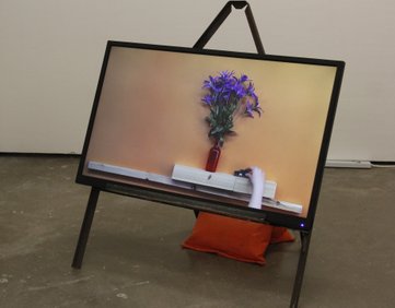 A modified road sign holding a landscape TV screen and an orange sandbag weighting it down. The TV is showing the film mantel piece